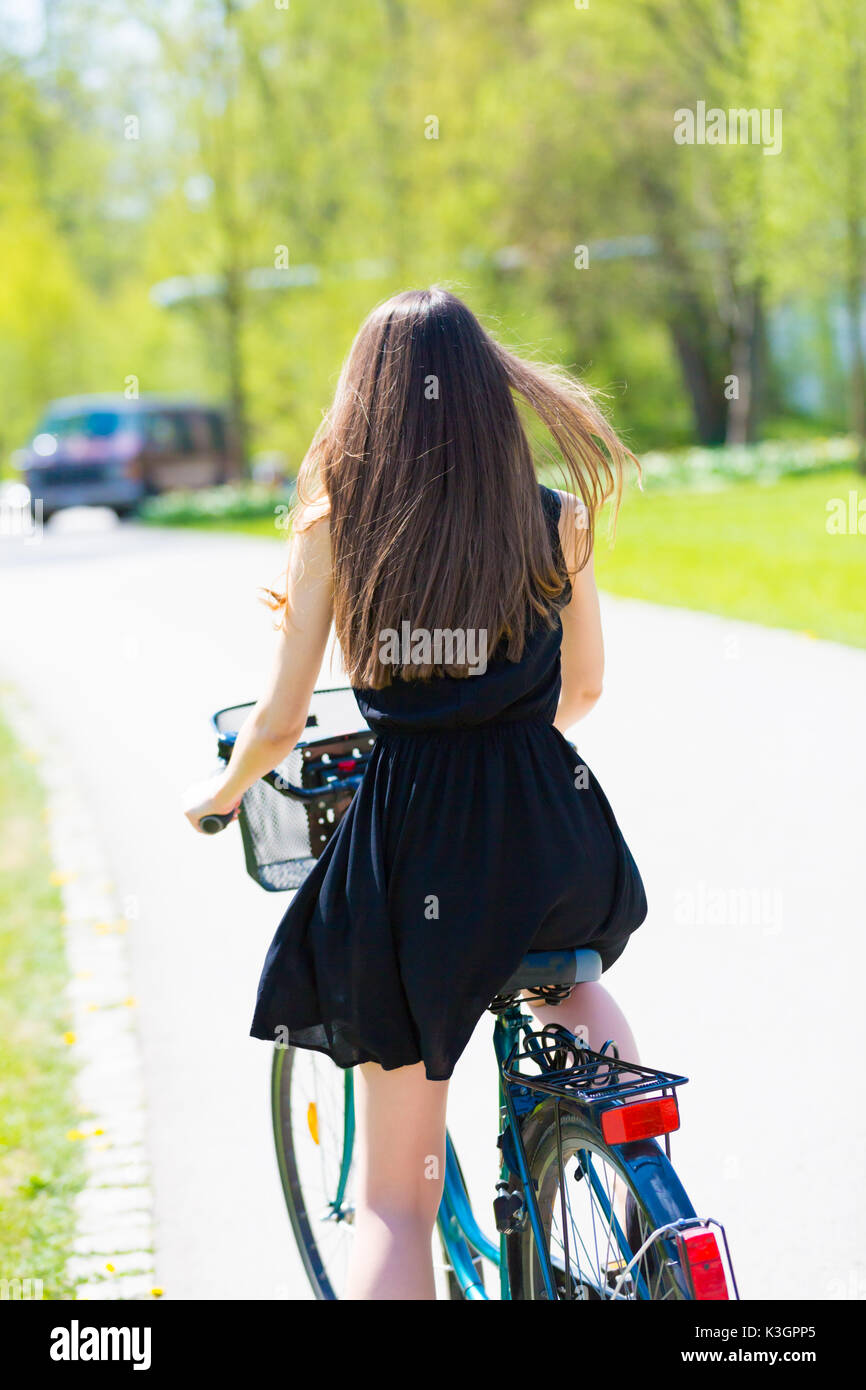 Rückansicht der Mädchen auf dem Fahrrad tragen am Schwarzen kurzen Kleid. Junge Frau Reiten entlang der Straße am grünen Frühling im Park. Sportlich junges Mädchen reiten ein Bic Stockfoto