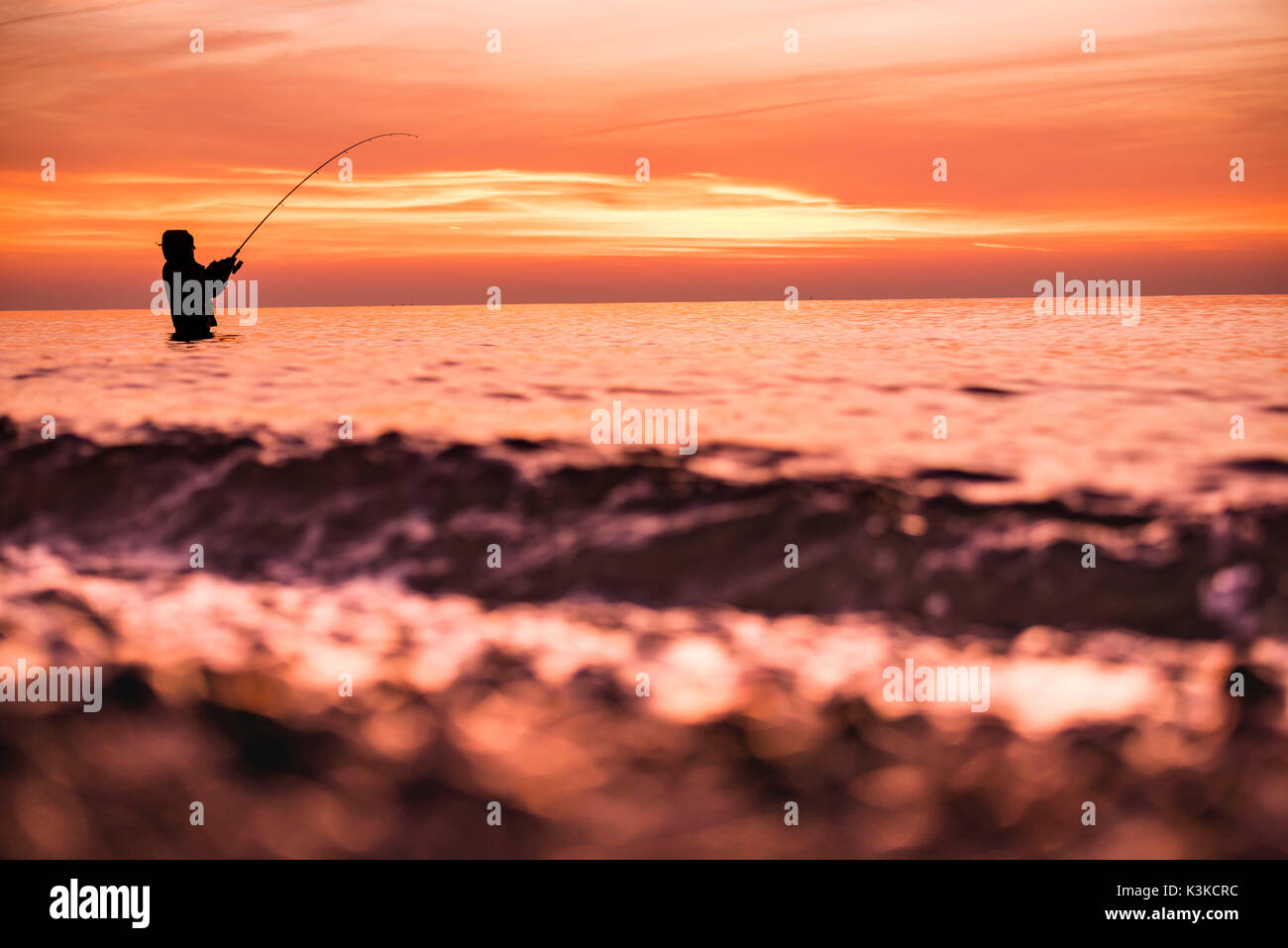Angler steht in das kalte Wasser der Ostsee bei Sonnenuntergang und würfelt eine Meerforelle. Im Vordergrund Wellen. Stockfoto