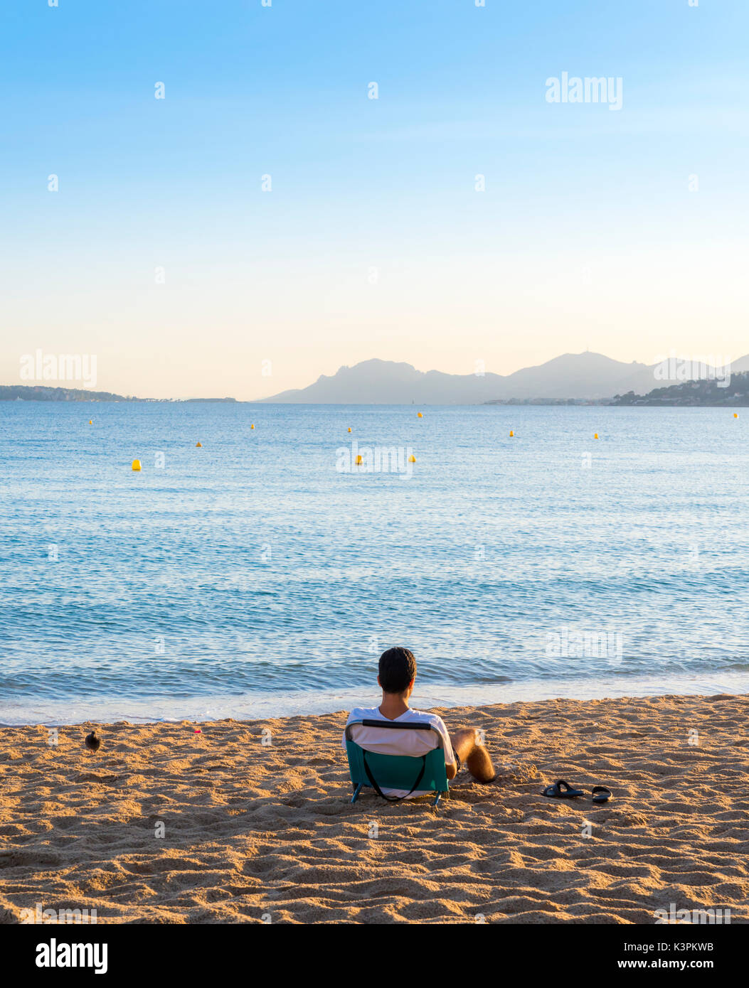 Ein junger Erwachsener sitzt auf einem leeren Strand Liege mit Blick auf eine Bucht in Cote d'Azur, Frankreich bei Sonnenuntergang Stockfoto