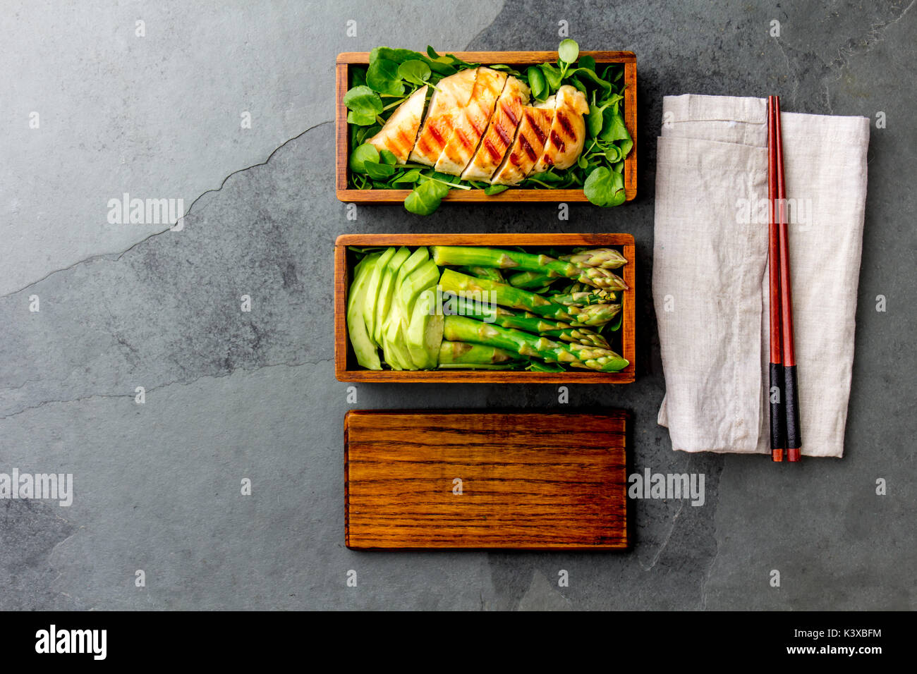 Gesundes Mittagessen in Holz- japanische Bento Box. Ausgewogene gegrillt chucken und Avocado mit Spargel und grüner Salat. Ansicht von oben, schiefergrau Bac Stockfoto