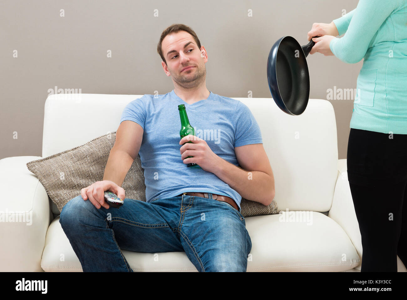 Frau schlagen mit Pan zu faul Mann sitzt auf einem Sofa Stockfoto