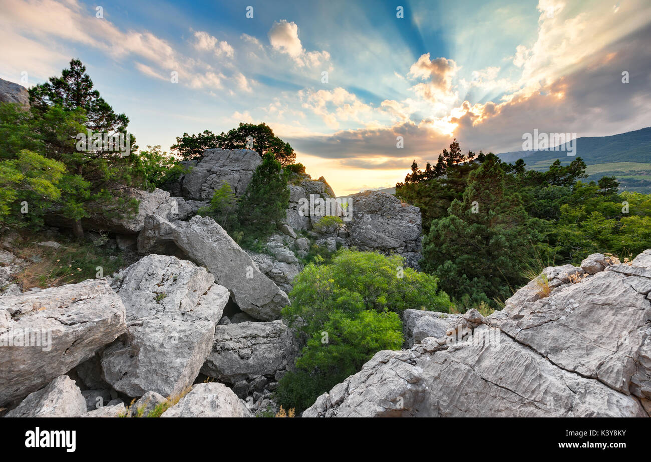 Haufen von Steinen auf dem Berg unter den Bäumen und Sträuchern im Hintergrund der untergehenden Sonne durch die Wolken Stockfoto