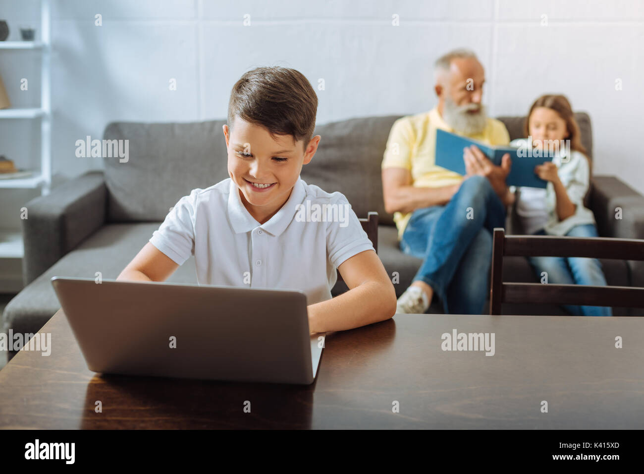 Junge spielt auf Laptop während Schwester und Großvater lesen Buch Stockfoto