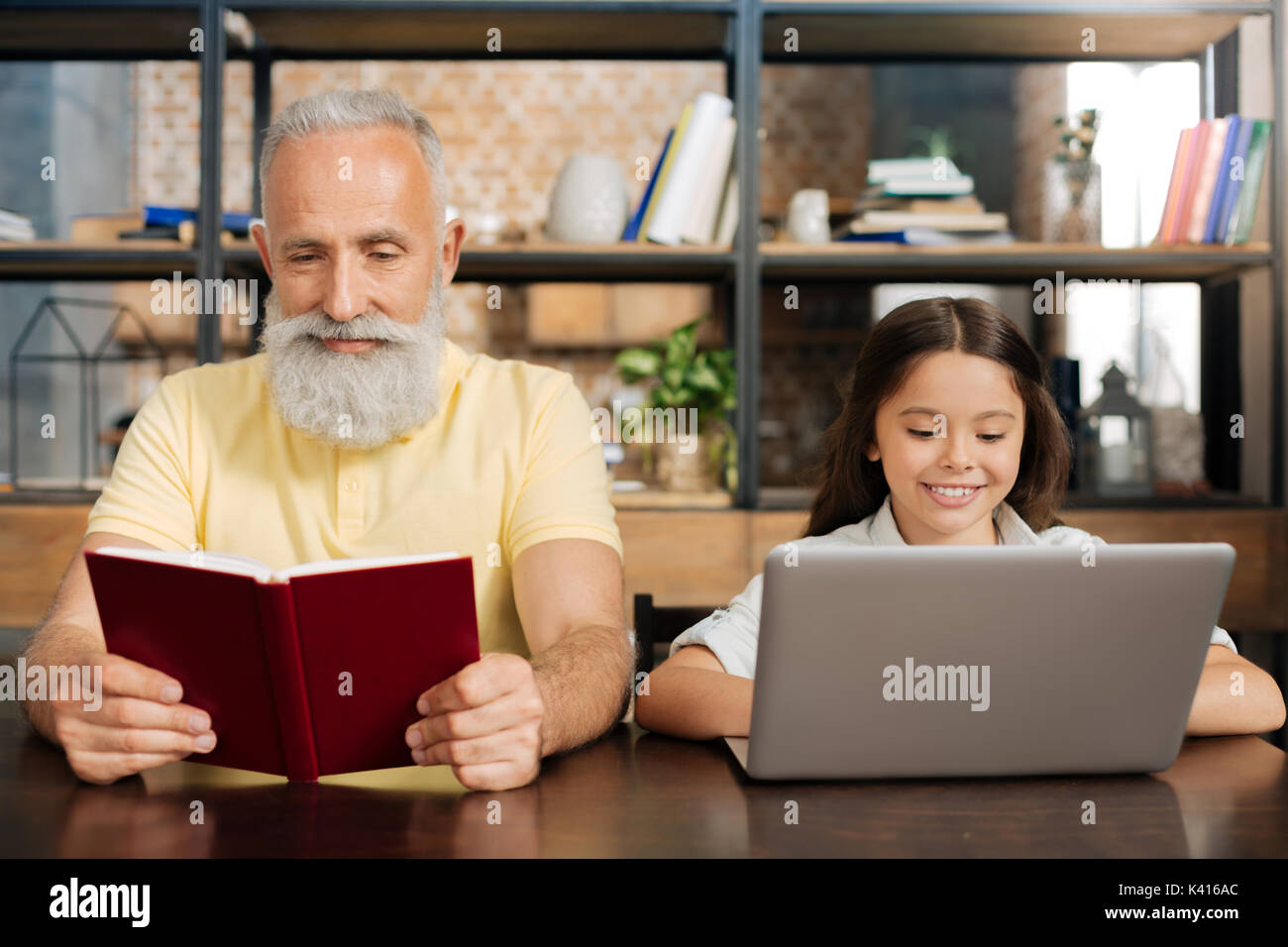 Angenehme Großvater lesen Buch, während seine Enkelin mit Laptop Stockfoto