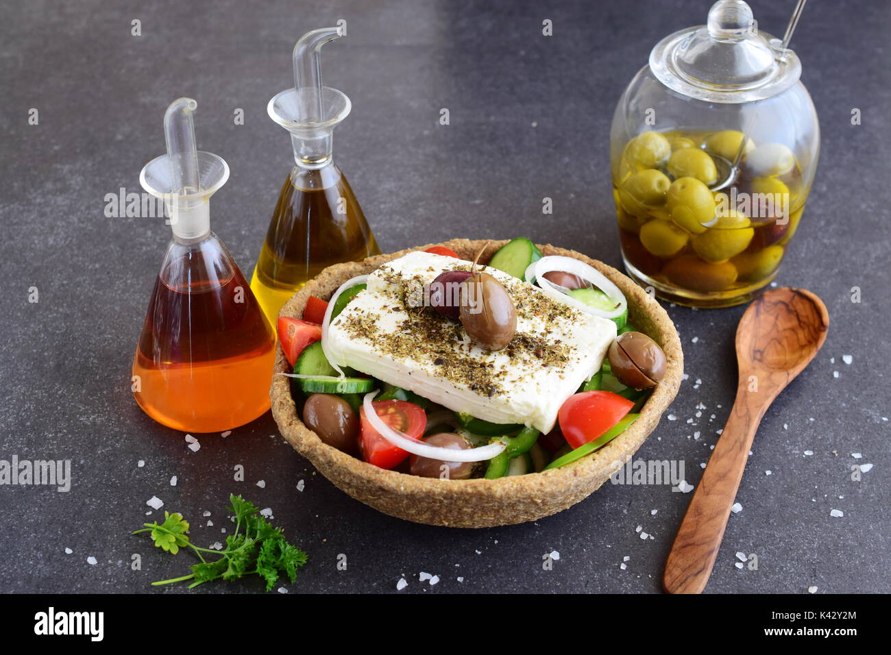 Traditioneller griechischer Salat mit Gurken, Zwiebeln, Tomaten, Paprika, Schafskäse, Olivenöl und Oregano in eine essbare Roggen Schüssel mit Gläsern mit Olivenöl, Essig und Oliven auf einer abstrakten Hintergrund Stockfoto