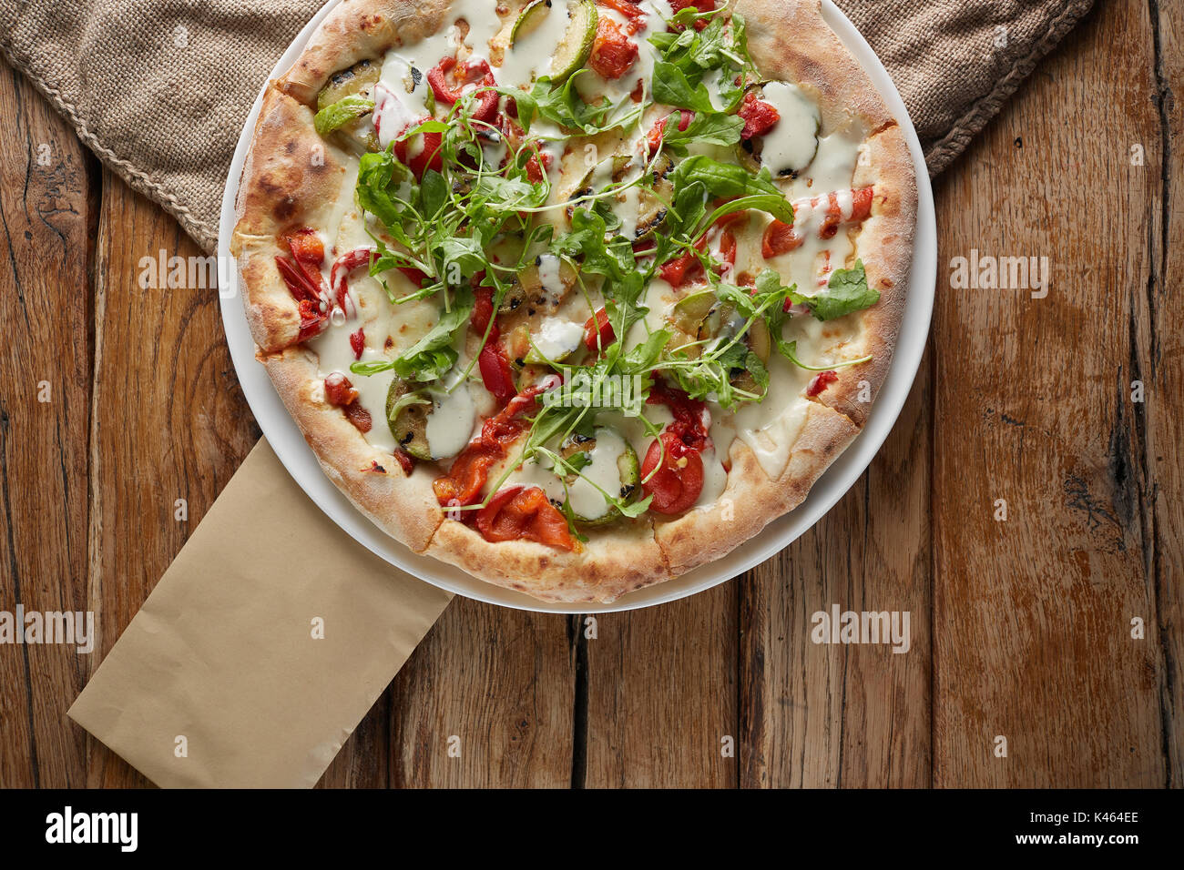 Oben Ansicht von Gerundeten große hausgemachte Pizza auf Weiß abgerundete Platte und Holz- Hintergrund. Leckere italienische Pizza mit Gemüse. Rote Paprika, Papier, Tomate Stockfoto