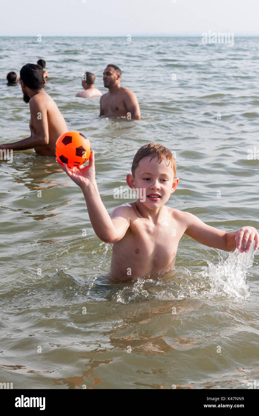 Ein achtjähriger Junge spielt mit einem orangefarbenen Ball in das Meer in Southend-on-Sea, Essex, Großbritannien Stockfoto