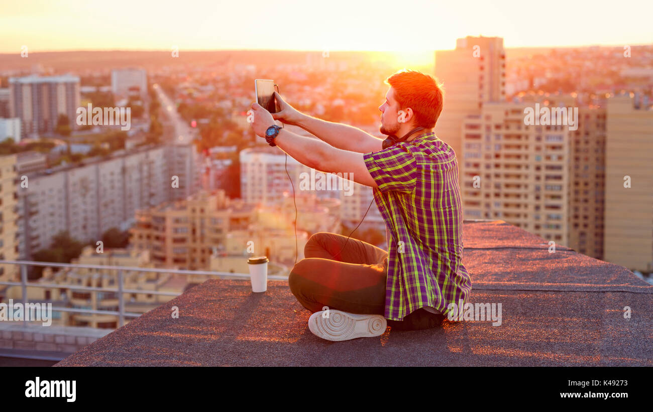 Hipster Mann Musik hören mit Kopfhörern auf dem Dach bei Sonnenuntergang. Er hält einen Tablet-PC in seinen Händen und nimmt Bilder auf die Stadt bei Sonnenuntergang Stockfoto