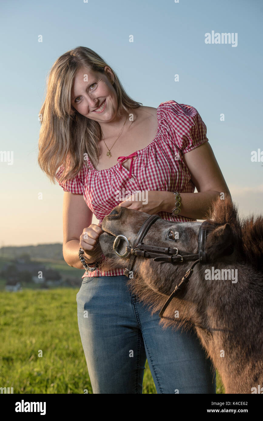 Lachende junge blonde Frau quält Brown Shetland Pony auf der Weide im weichen Licht des Sonnenuntergangs Stockfoto