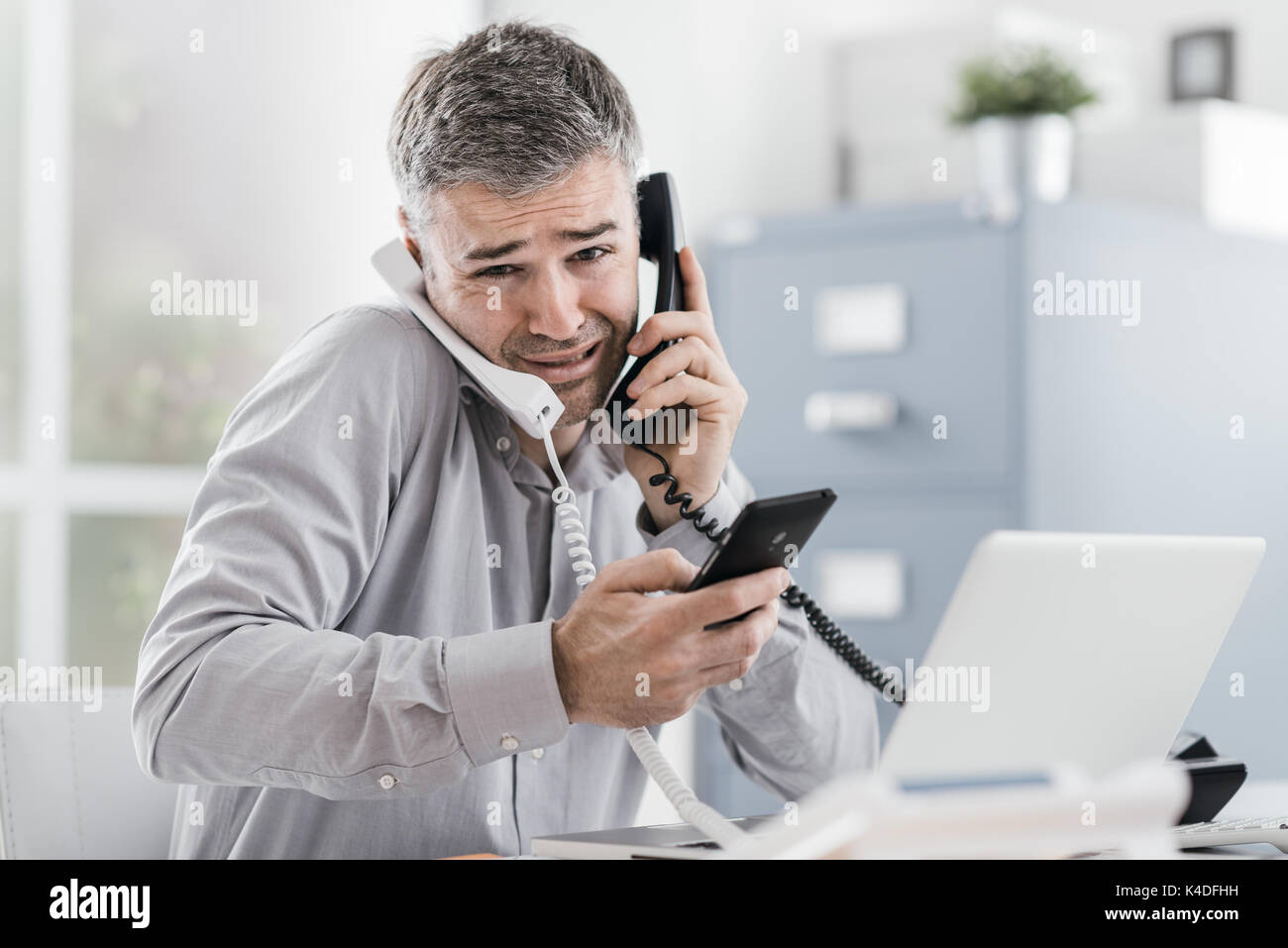 Verzweifelt Geschäftsmanns, der in seinem Büro und in mehrere Anrufe betonte, er hält zwei Mobilteile und ein Handy, Business Management con Stockfoto
