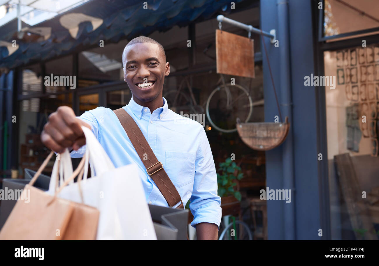 Porträt eines eleganten jungen afrikanischen Mann und lächelnd bis Einkaufstaschen beim Stehen vor einem Geschäft in der Stadt Stockfoto
