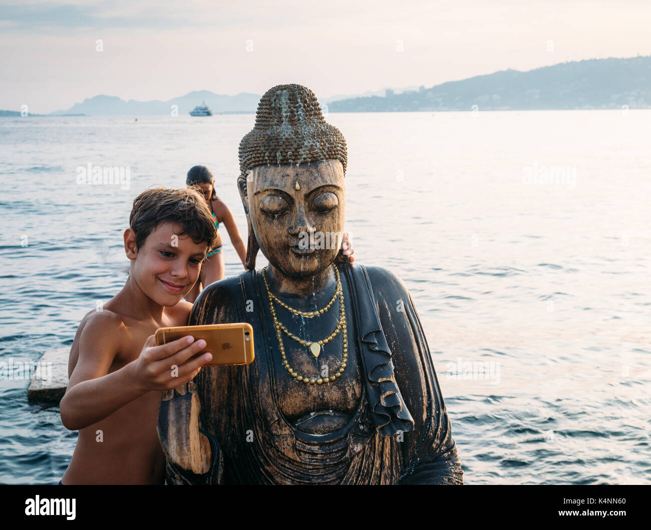 Ein kleiner Junge nimmt eine selfie mit einer Statue des Buddha auf einem Strand in Juan-les-Pins, Frankreich Stockfoto