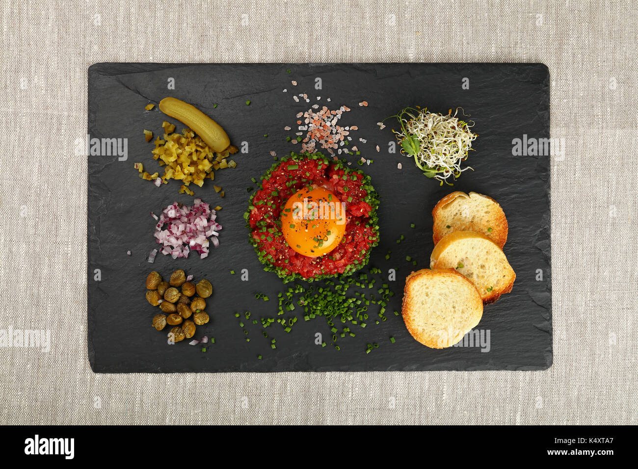 Eine Portion rohes Hackfleisch Fleisch Steak Tartar mit Eigelb, Grün, Schnittlauch, Zwiebeln, Gurken, Kapern, Salz und Toast am schwarzen Schiefer Board auf Tisch Stockfoto