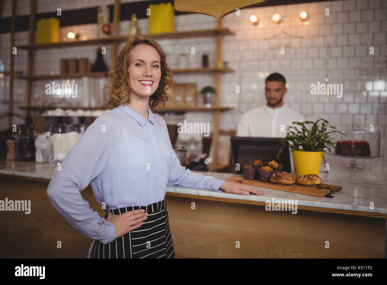 Portrait von lächelnden Kellnerin stehen mit Hand auf der Hüfte durch Zähler bei Coffee Shop Stockfoto