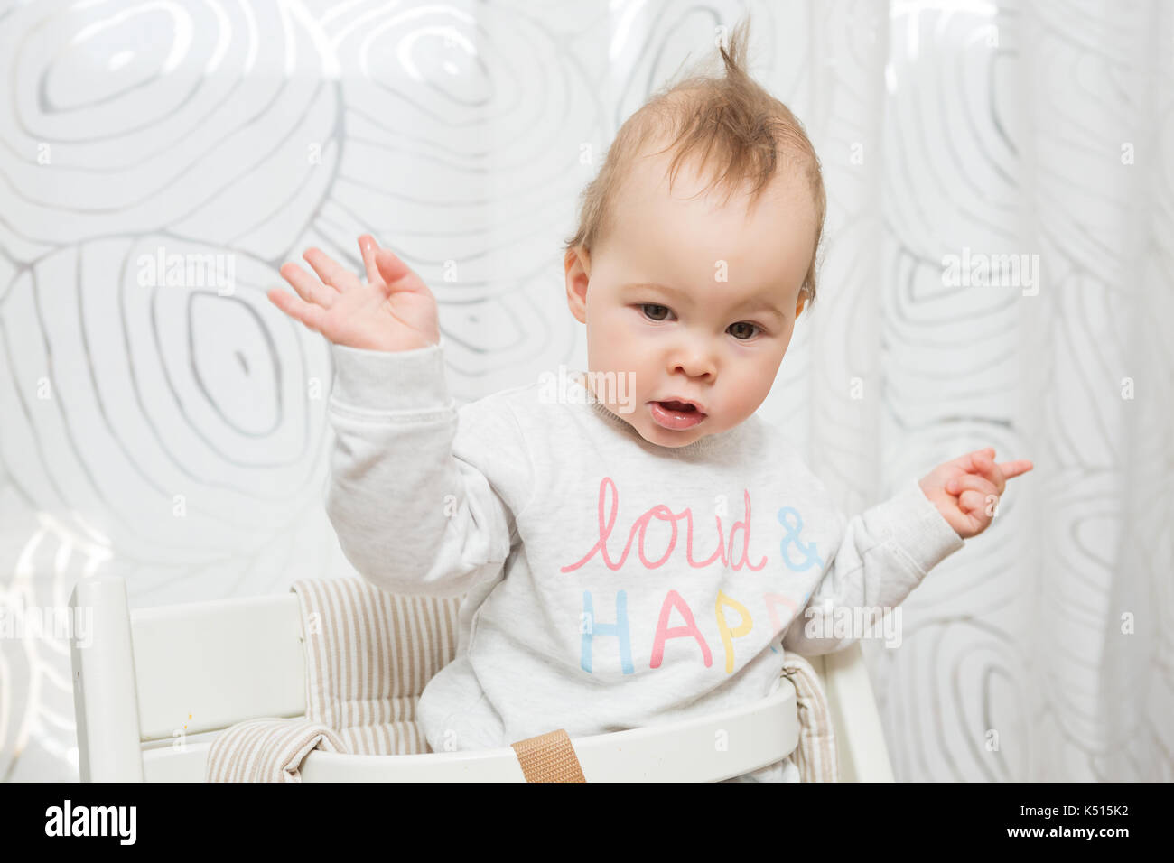 Elf Monate alten Baby Mädchen in einem Hocker, lustige Gesichter in die Kamera sitzen, heben Sie beide Hände in der Luft Stockfoto