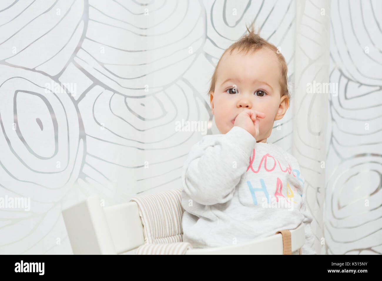 Elf Monate alten Baby Mädchen sitzen auf einem Stuhl, lustige Gesichter in die Kamera und ihre Finger im Mund Stockfoto