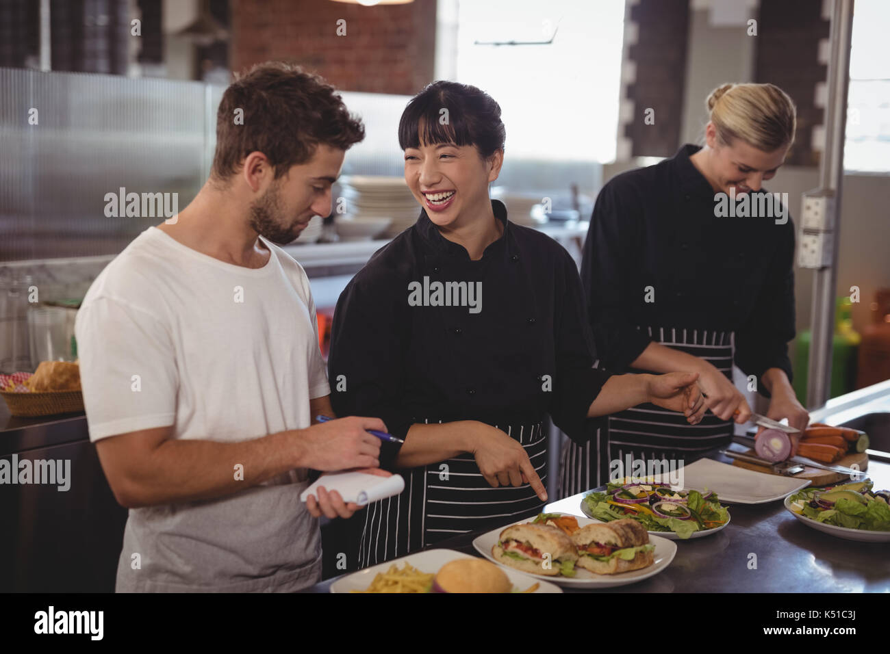 Fröhlicher Koch an Nahrungsmittel, die von den weiblichen Kollegen im Café Küche, Kellner Stockfoto