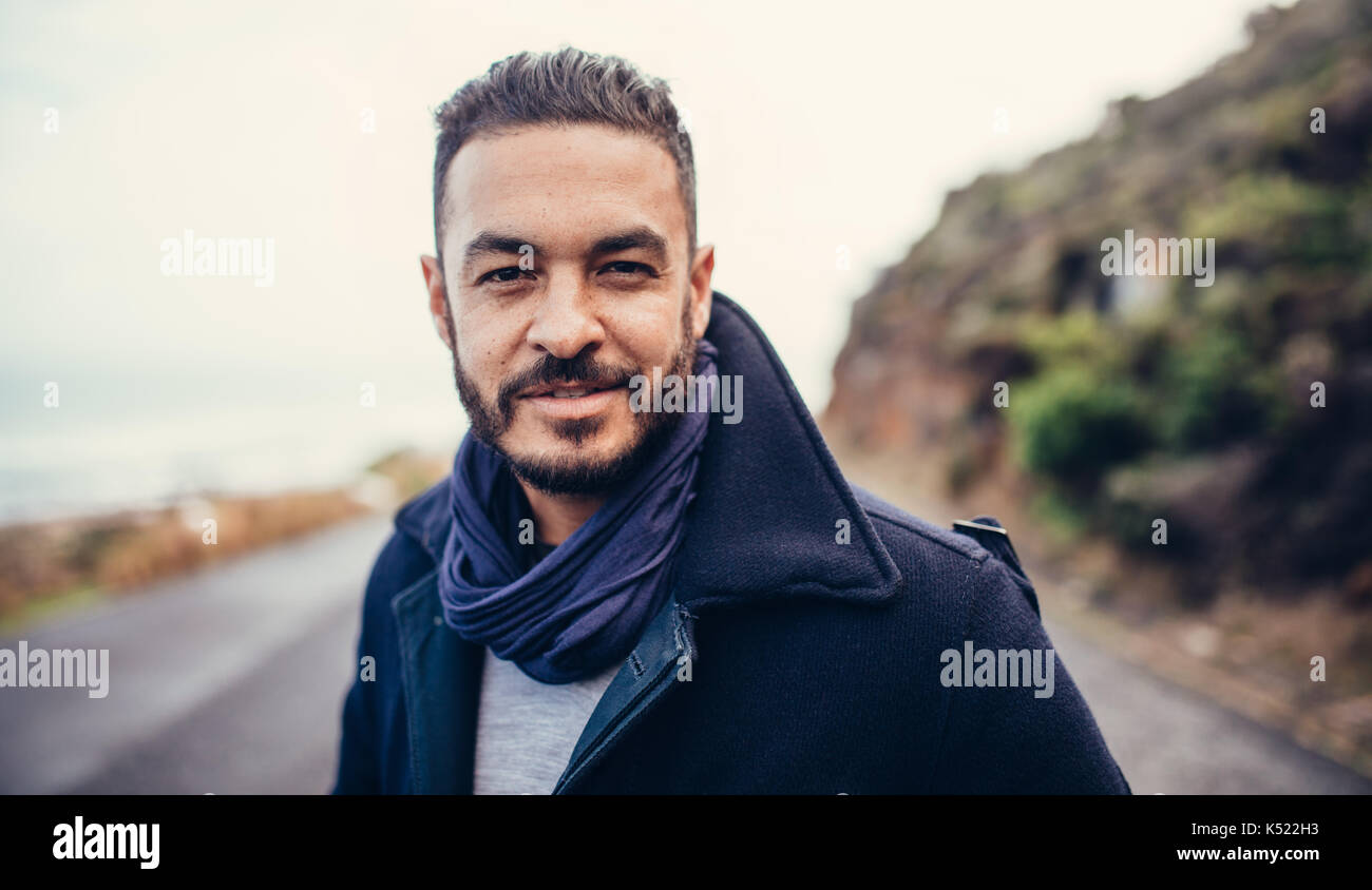 Hübscher junger Mann auf der Straße mit Bergen im Hintergrund. Mann im Winter Kleidung auf Kamera. Stockfoto