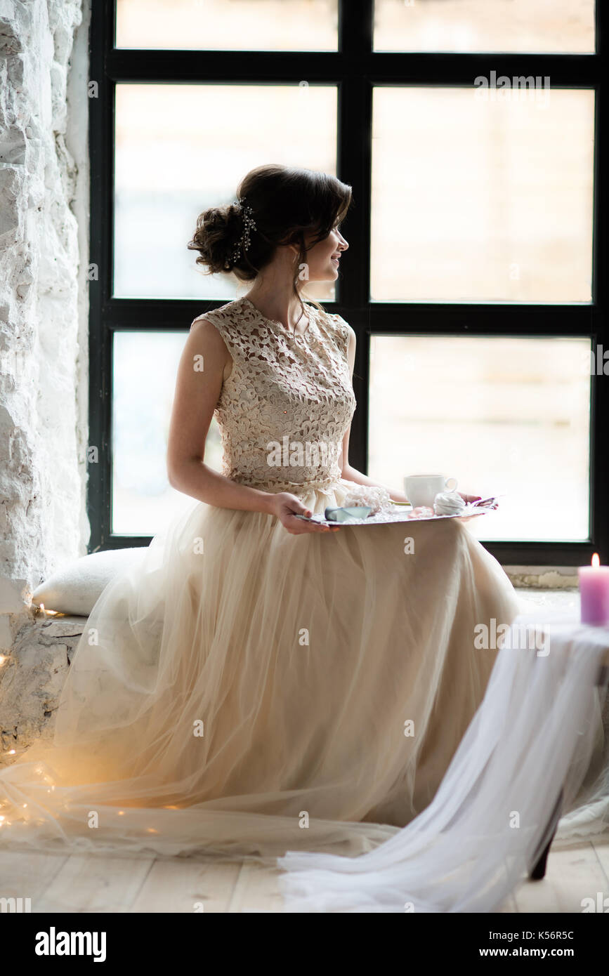 Junge wunderschöne Frau tragen ärmelloses Abendkleid. Sie wartet auf ihren Freund sitzen vor dem Fenster mit Tablett mit Süßigkeiten und Kaffee. Stockfoto