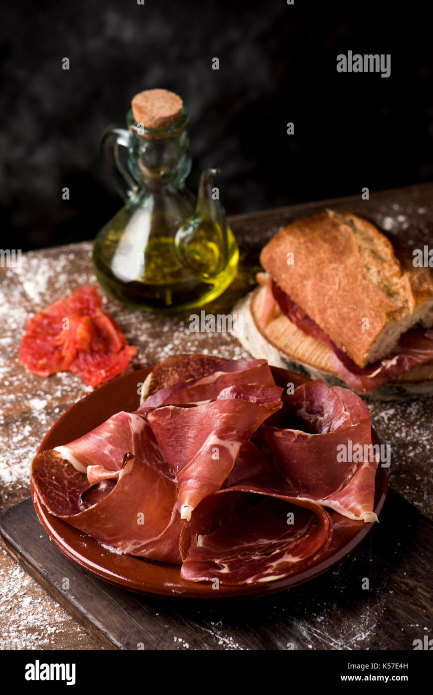 Eine Platte mit einigen Scheiben Serrano Schinken auf einem rustikalen Holztisch, neben einem cruet mit Olivenöl und ein Sandwich von typisch katalanischen Pa amb Tomaquet, br Stockfoto