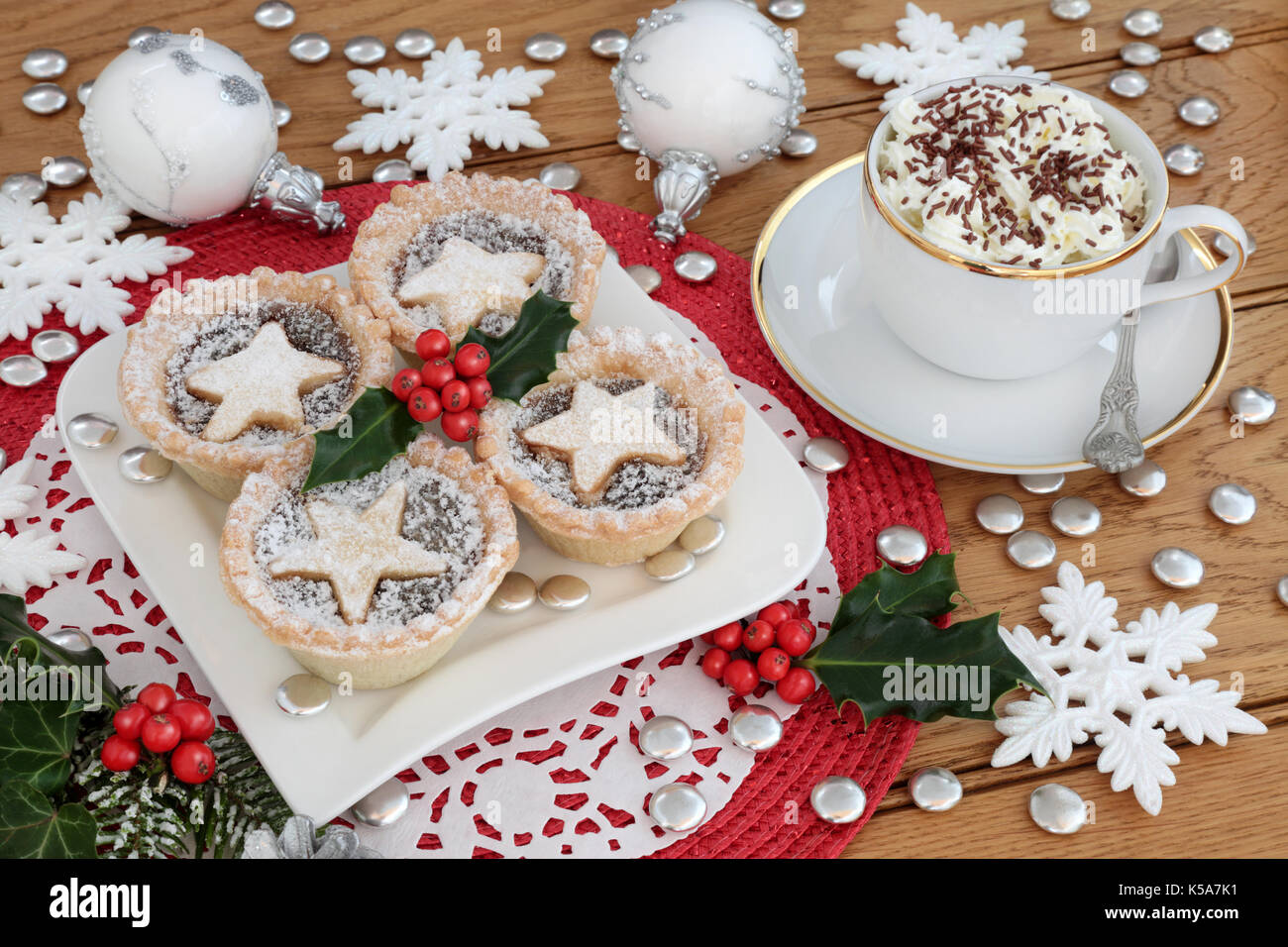 Weihnachten Mince Pies auf einem Teller mit heiße Schokolade trinken, Holly und Christbaumkugel Dekorationen auf einem weißen dolie auf einem roten Unterlage auf Eiche Hintergrund. Stockfoto