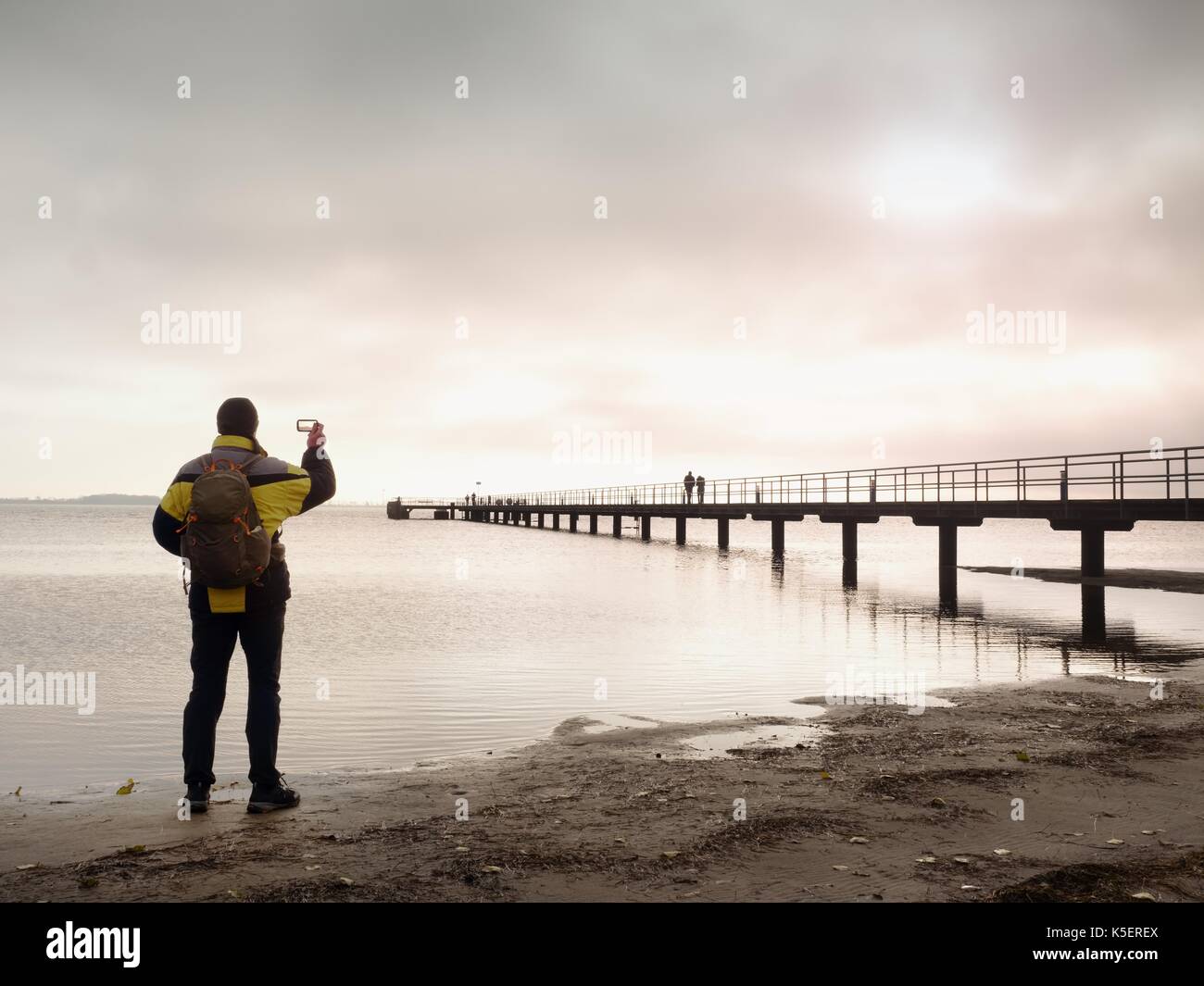 Mann Wanderer mit Rucksack stand alone auf schmutzigen Sandstrand und romantischen farbenfrohen Sonnenaufgang über hölzerne Seebrücke. Vintage Style getönten Wirkung Stockfoto