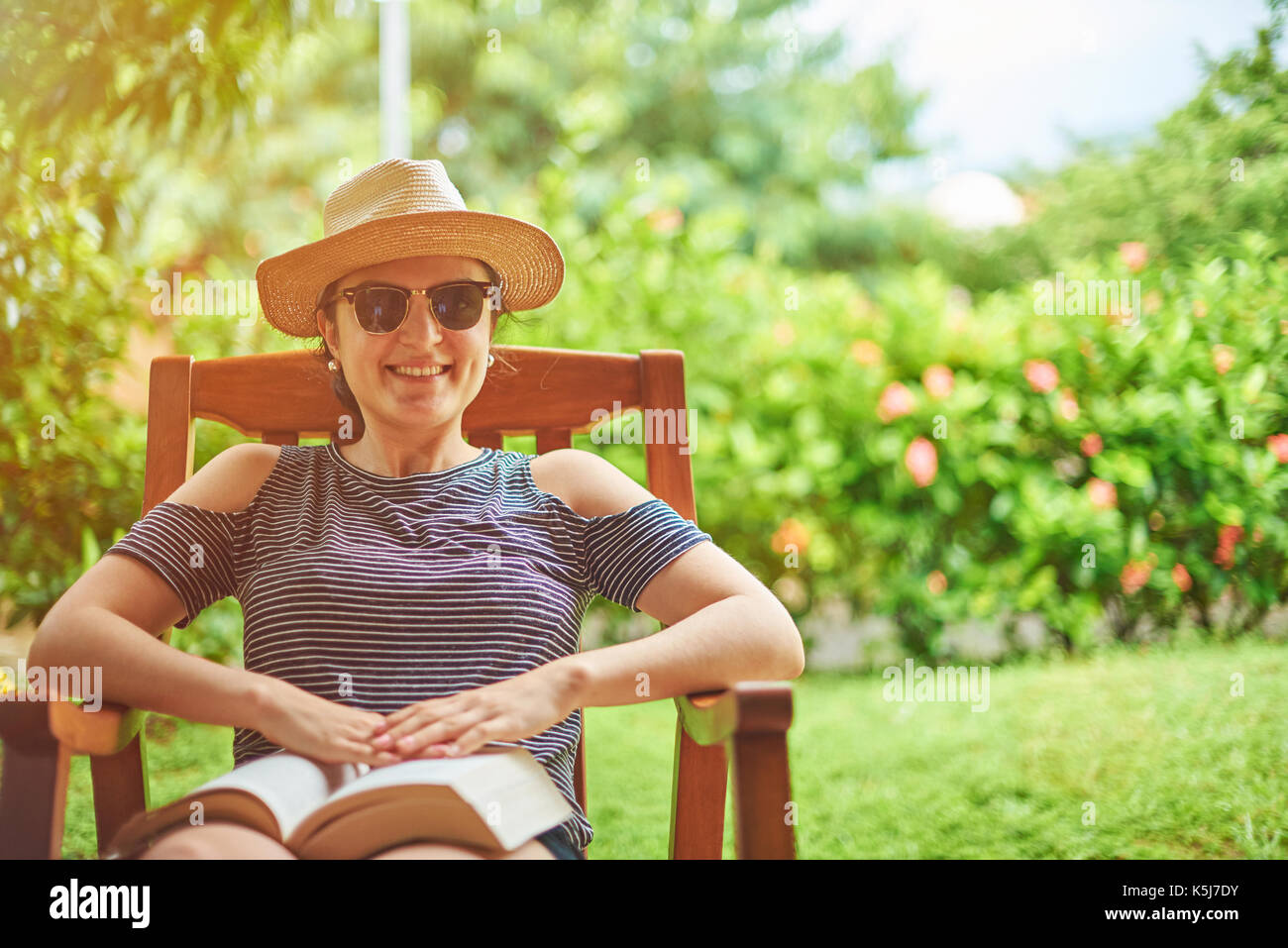 Lächelnde Frau mit Buch an sonnigen, grünen Garten Terrasse Stockfoto