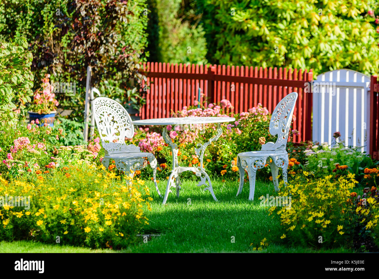Weißes Metall Gartenmöbel unter den Blumen auf einer Wiese. Lattenzaun im Hintergrund. Stockfoto