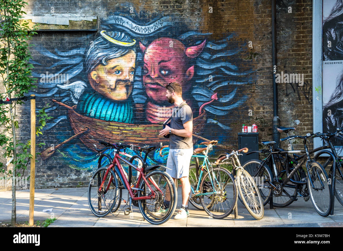 Ein Mann sammelt auf seinem Fahrrad von einem Fahrrad Rack auf Castlehaven Straße in Camden Town, London. Ein wandbild wird auf die Wand im Hintergrund. Stockfoto