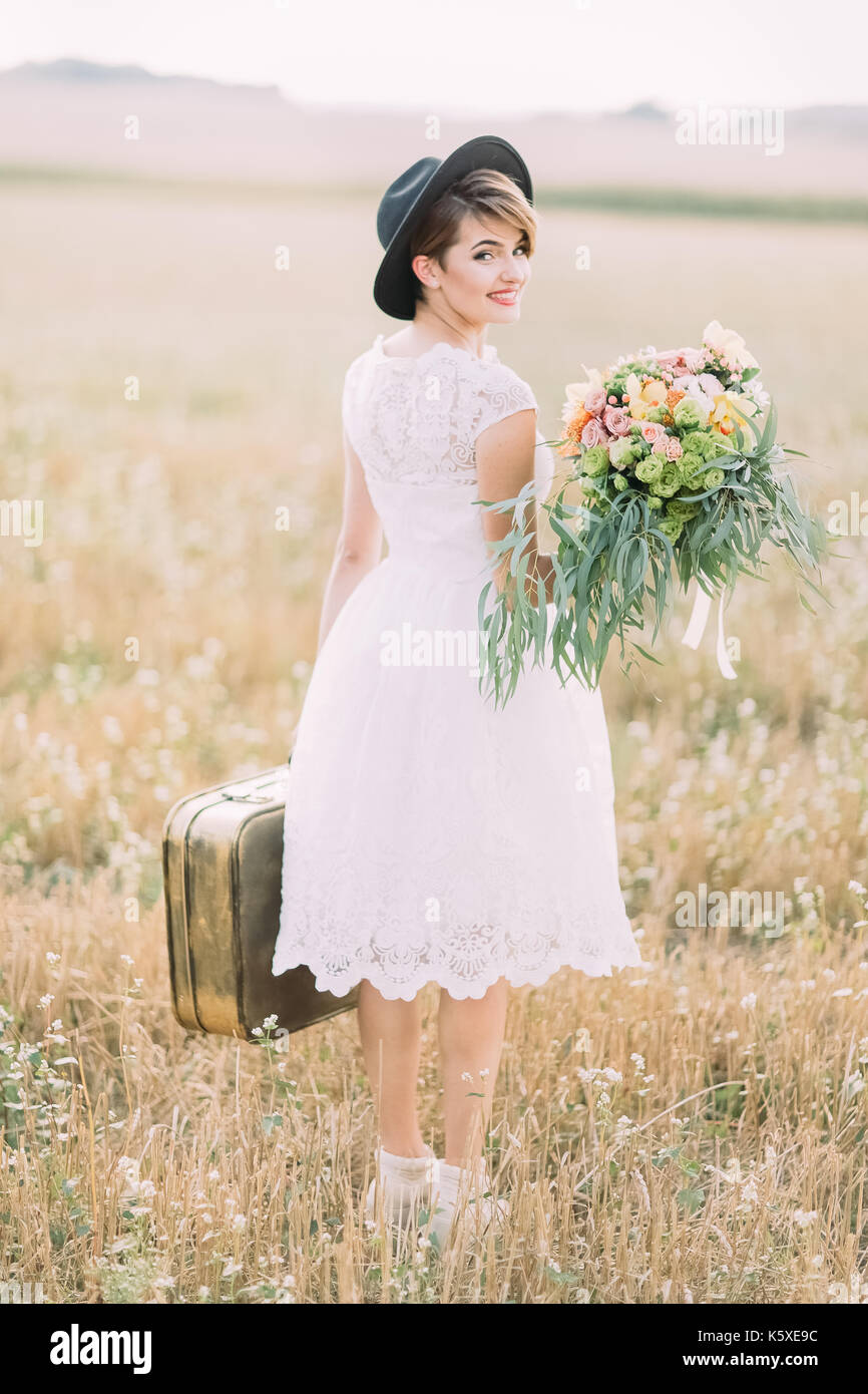 Die Rückansicht der Vintage gekleidete Braut die Hochzeit Bouquet, die altmodische Koffer und lächelnd in die Kamera im Hintergrund des Feldes. Stockfoto