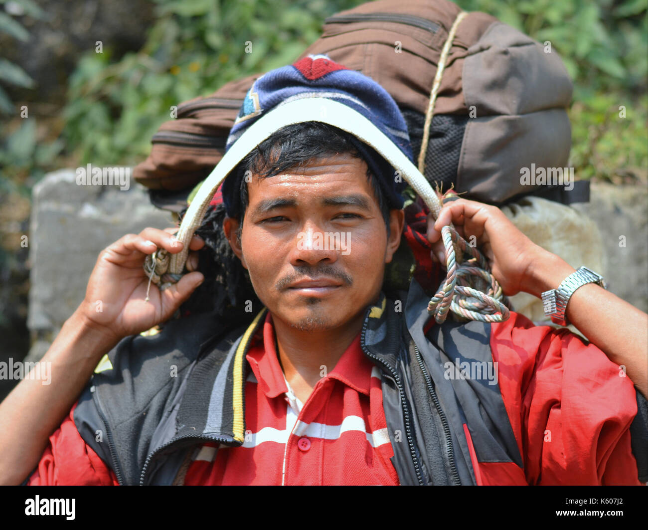 Der junge nepalische Bergportier (Sherpa) mit einer schweren Last macht eine kurze Pause auf einer Steinbank und posiert für die Kamera. Stockfoto