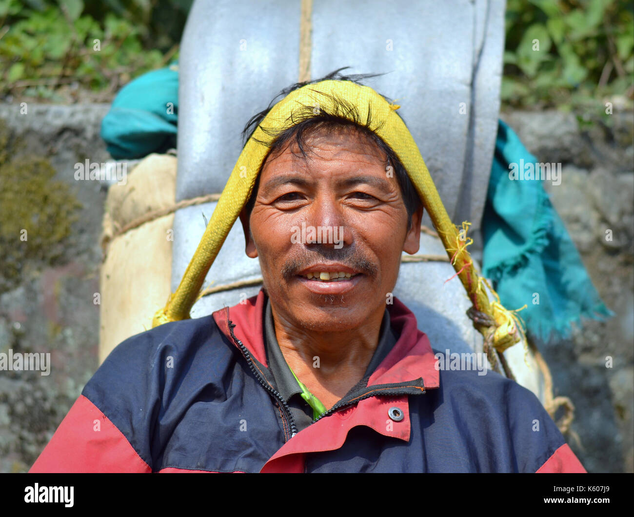 Der nepalische Bergportier (Sherpa) mit einer schweren Last macht eine kurze Pause auf einer Steinbank und lächelt für die Kamera. Stockfoto