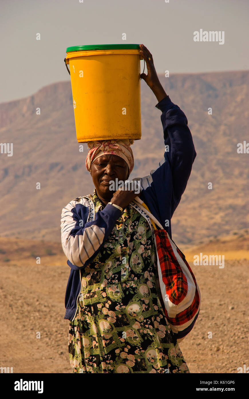 Afrikanische Frau, die einen Eimer voller Wasser o seinen Kopf an der Fernbedienung und Land im Norden von Namibia in der Nähe von Palmwag Stockfoto
