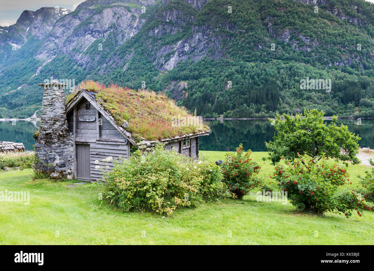 Alte märchenhafte Haus aus Holz mit Kamin aus gestapelten Schiefer und ein Dach mit Pflanzen und Bäume an einem Fjord in Norwegen mit Garten mit roten Blumen ein Stockfoto