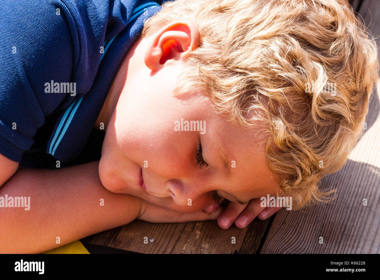 Kind, kleine blonde lockige schwarzhaarige Junge, in der Nähe des Gesichtes, das so tut, schlafend, den Kopf auf den Händen ruht während der Verlegung auf die Holzterrasse. Stockfoto