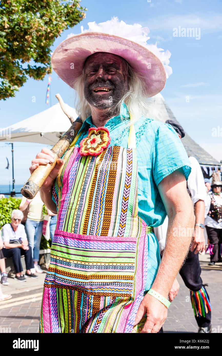 Portrait der männlichen 'Idiot' von Dead Horse Morris Dancers. Augenkontakt, geschwärzte Gesicht, ein Lächeln auf den Lippen. Trägt Damen rosa Hut, blaues Kleid und bunten Schürze. Stockfoto