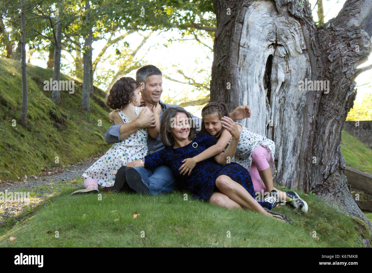 Eltern, die ihre beiden Töchter mit der Nachbearbeitung in einem Sommer Urlaub Hug unter einem Baum Stockfoto