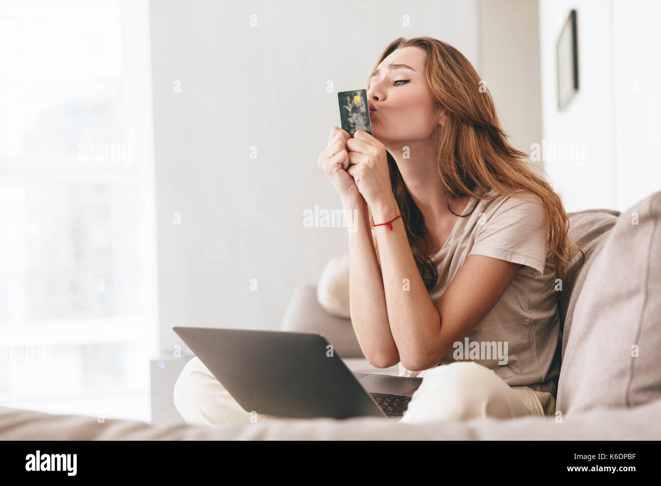 Lustige junge Frau in der beiläufigen Kuss ihre Kreditkarte beim Sitzen mit Laptop auf dem Sofa zu Hause Stockfoto