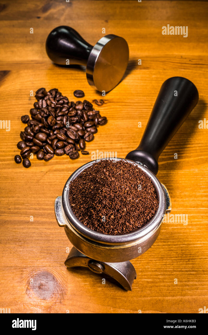 Eine Handvoll Kaffeebohnen, neben den gemahlenen Kaffee in eine Espressomaschine portafilter Korb aus Stahl und ein stampfer, alle sitzen auf einem mit Pinien Arbeitsplatte. Stockfoto