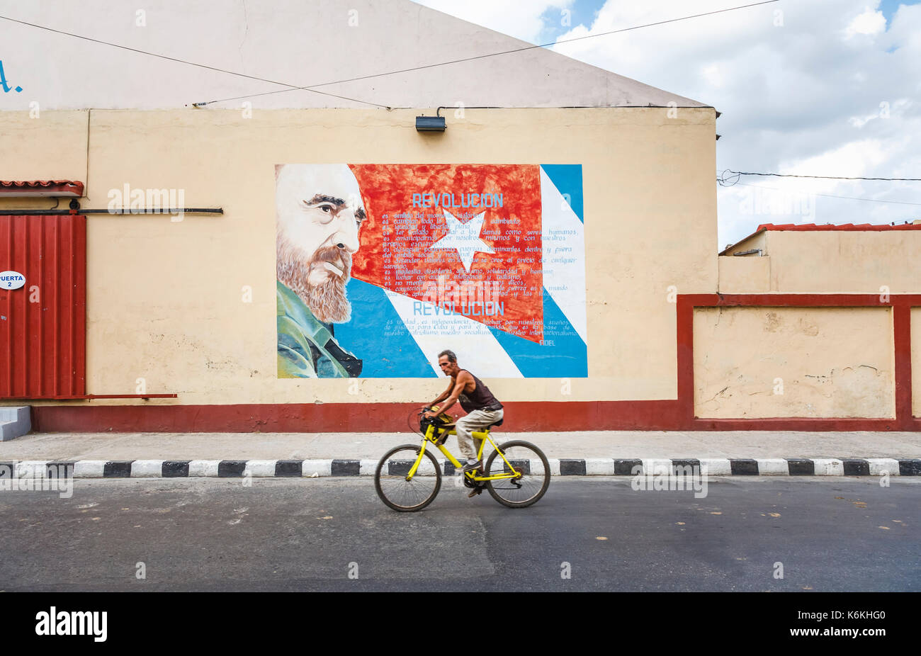 Einheimischer Mann, der mit dem Fahrrad an einer Straßenmauer vorbeifährt, die Fidel Castro und die kubanische Flagge zeigt, Cienfuegos, eine Stadt an der Südküste Kubas Stockfoto