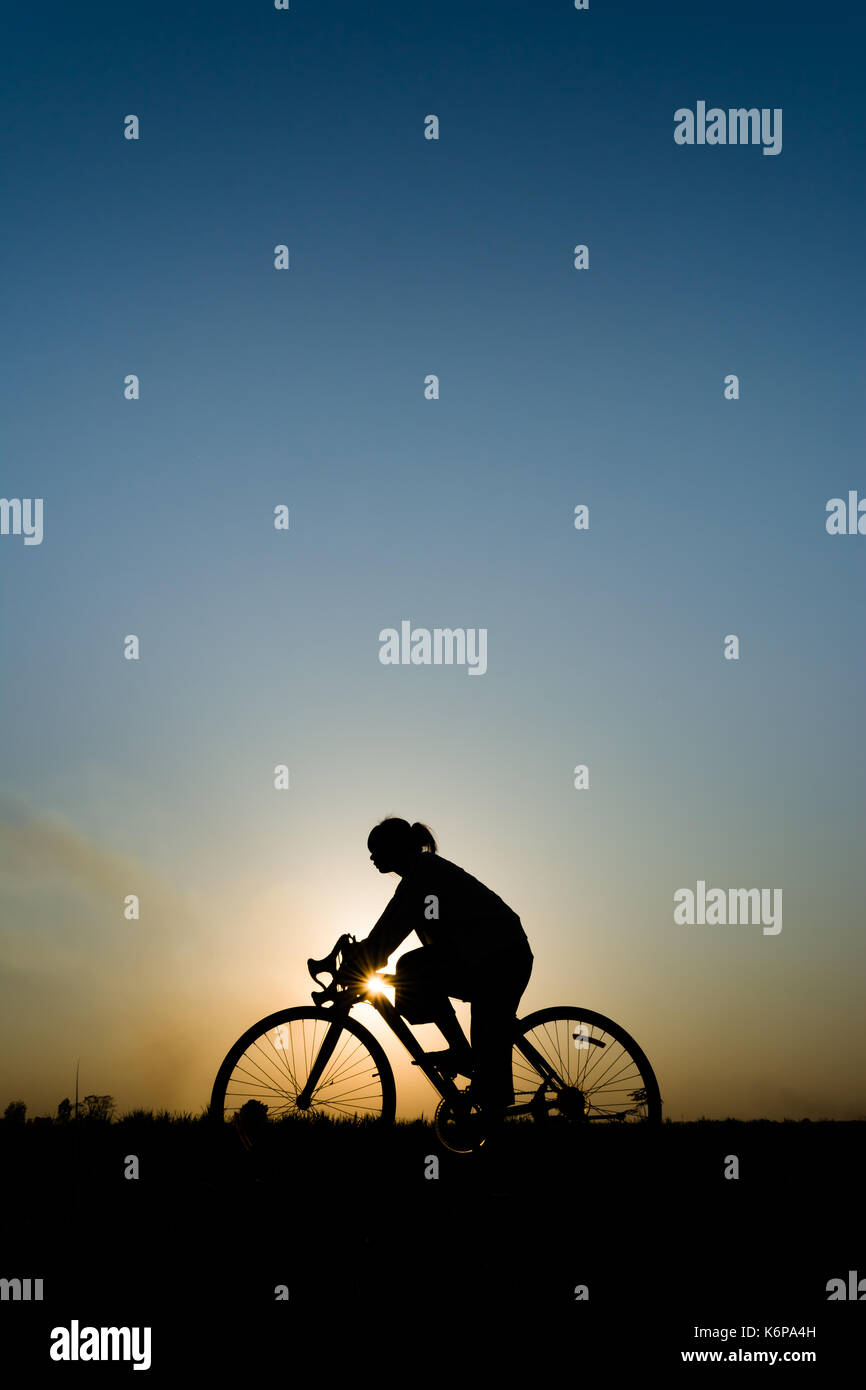 Silhouette der jungen Frau Radfahrer auf Sonnenuntergang Himmel mit Reiten entlang der Wiese am gelben Abend Horizont Meer gelber Sonnenuntergang Himmel Hintergrund im Freien Stockfoto
