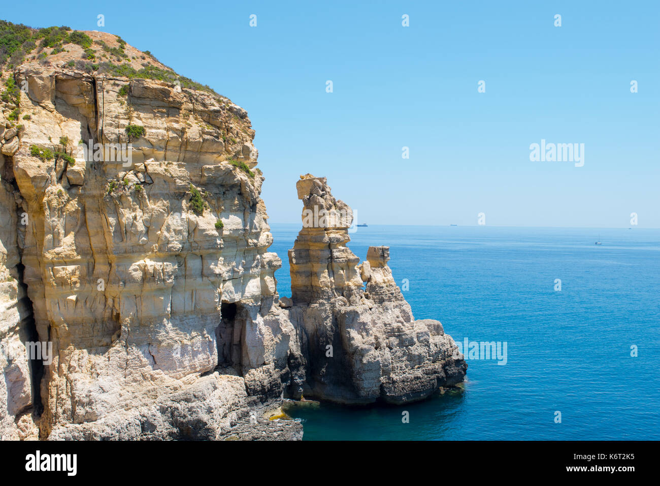 Ein Kalkstein Klippe mit einer kleinen Höhle und zwei short stacks, entlang der maltesischen Küste gefunden. Das Meer ist ruhig mit einem Handelsschiff Ferne. Stockfoto