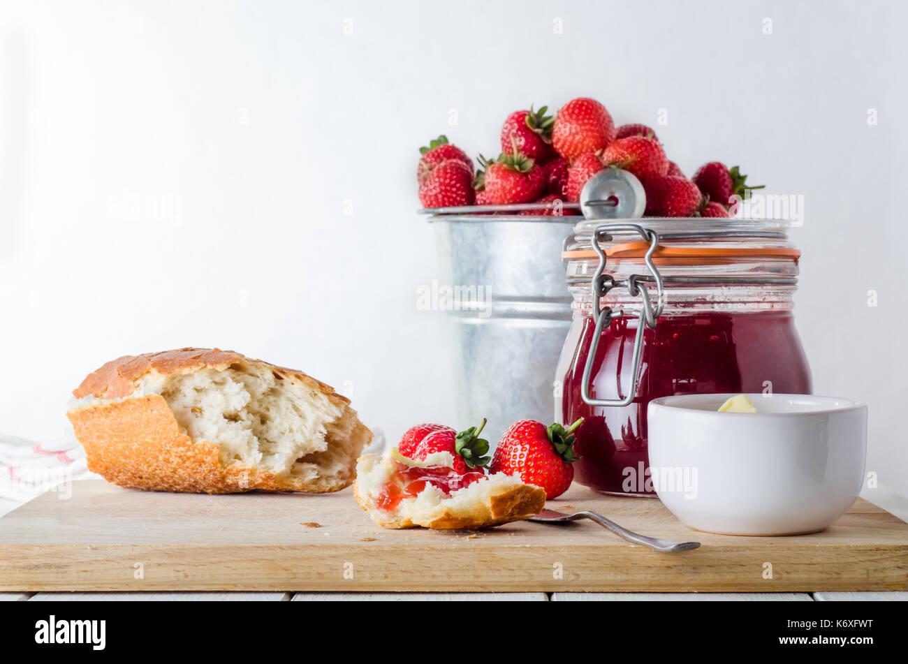 Eine Küche Tisch Szene von frisch gemachte Erdbeermarmelade in Storage jar mit einer Dose Eimer von Erdbeeren im Hintergrund; Brot, Butter und Erdbeeren Stockfoto