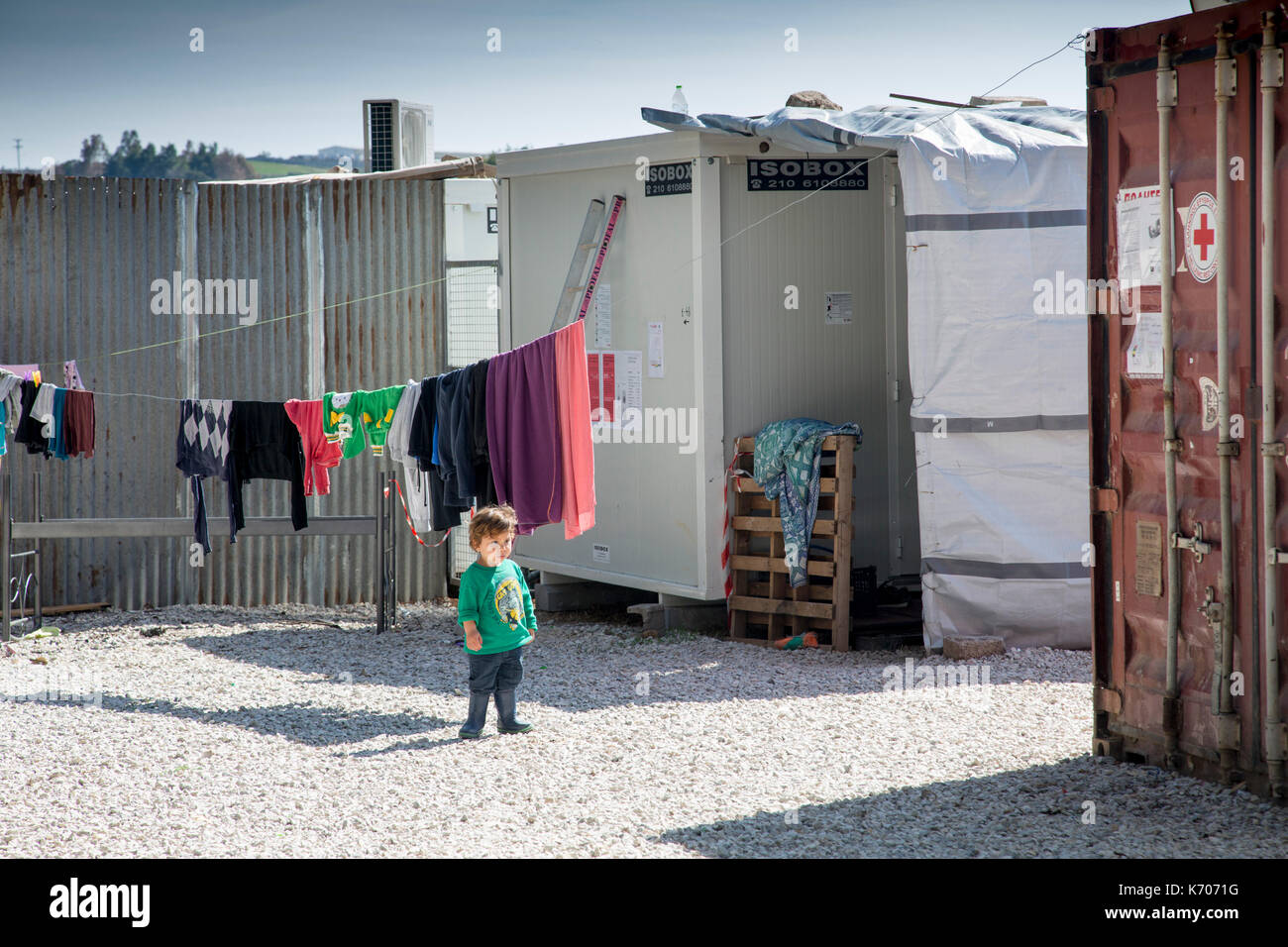 Auf dem Kies zwischen vorgefertigten Einheiten, in denen Flüchtlinge aus Syrien untergebracht sind, untersucht ein einjähriges Kind das Flüchtlingslager Ritsona. Vordergrund – ein Container des Roten Kreuzes. Stockfoto