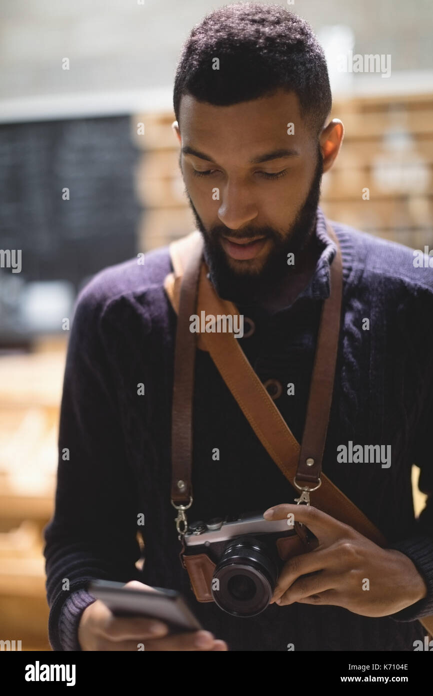 Der junge Mann mit der Kamera mit Handy während im Cafe stehend Stockfoto