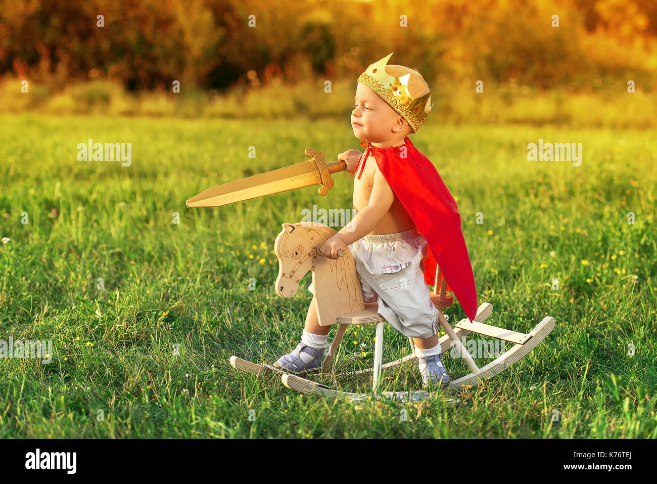 Der König das Kind rittlings auf einem Pferd spielt mit einem Schwert. Kleine Prinz mit einer Krone auf dem Kopf und in einem roten Regenmantel. Stockfoto