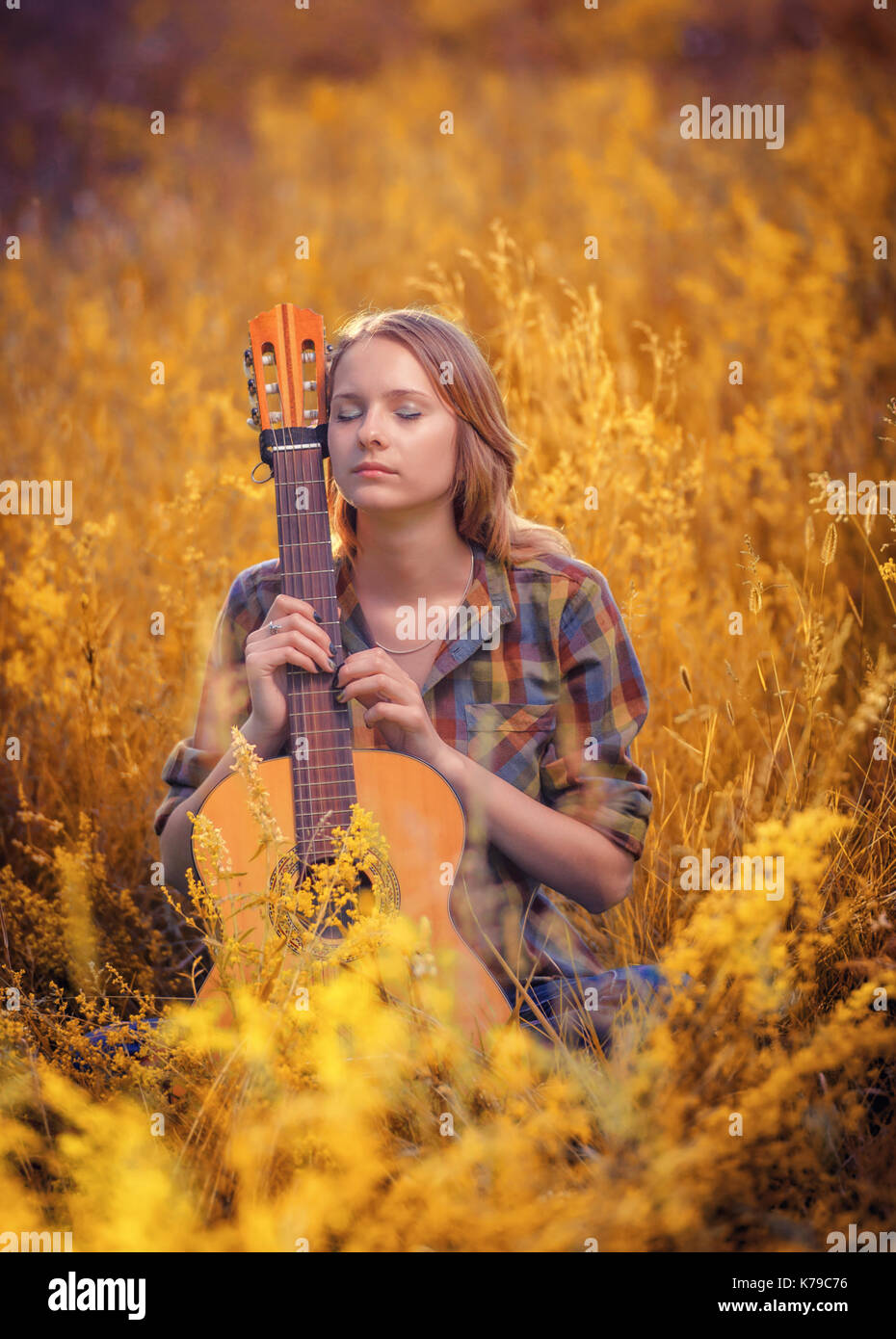 Junge schöne Mädchen mit geschlossenen Augen sitzt in einem Feld in eine akustische Gitarre. Romantisches herbst Stimmung bei Sonnenuntergang Stockfoto