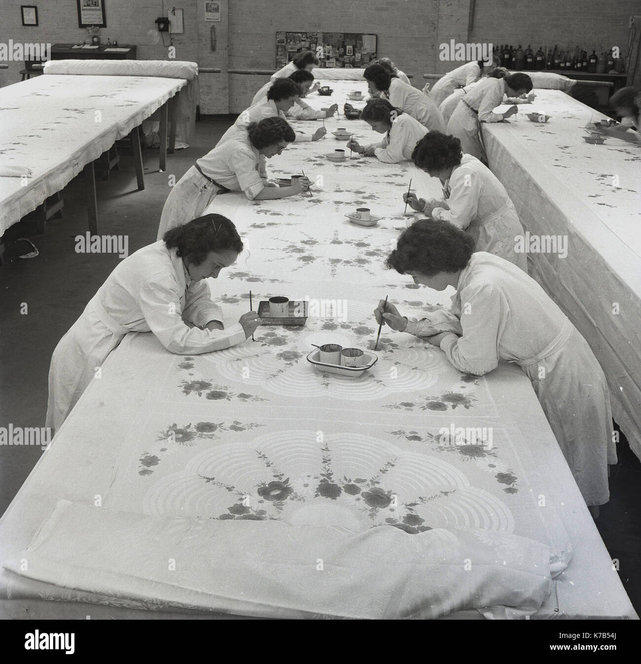 Eine historische Gruppe von Frauen aus den 1950er-Jahren, die auf langen Tischen mit Pinsel Entwürfe auf gewebte Leinentücher gemalt haben, Nordirland. Irisches Leinen ist ein Naturstoff mit langer Tradition, der aus der Leinpflanze (Unkraut) hergestellt wird, die natürlich auf argem Land und bei Regen wächst. Stockfoto