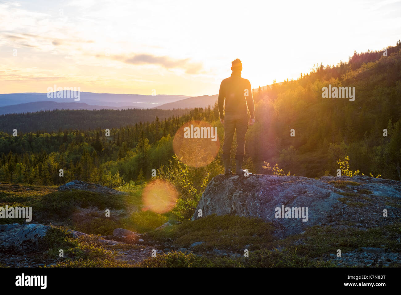 Junger Mann steht auf einem großen Stein und blickt in den Sonnenuntergang inmitten der Landschaft Stockfoto