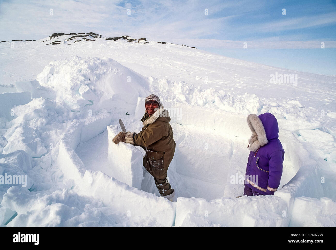 Inuit ältester Mann, Mitte der 60er Jahre, in der modernen Winter Kleidung, Schnitte Eisblöcke mit Messer und schneidet sie während Gebäude im traditionellen Iglu. Seine 4-jährige Enkelin Uhren. Stockfoto